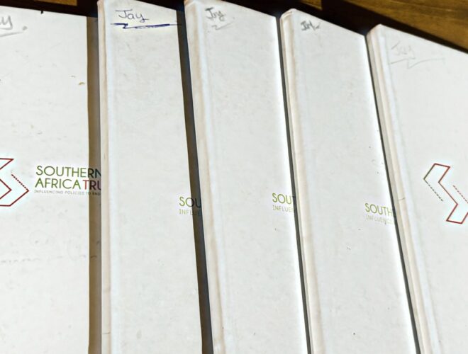 Five white Southern Africa Trust hardcover notebooks fanned out on a wooden table, each one marked "Jay" by hand in the top corner, the covers worn from years of use.