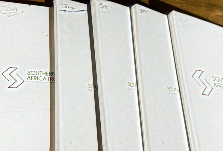 Five white Southern Africa Trust hardcover notebooks fanned out on a wooden table, each one marked "Jay" by hand in the top corner, the covers worn from years of use.