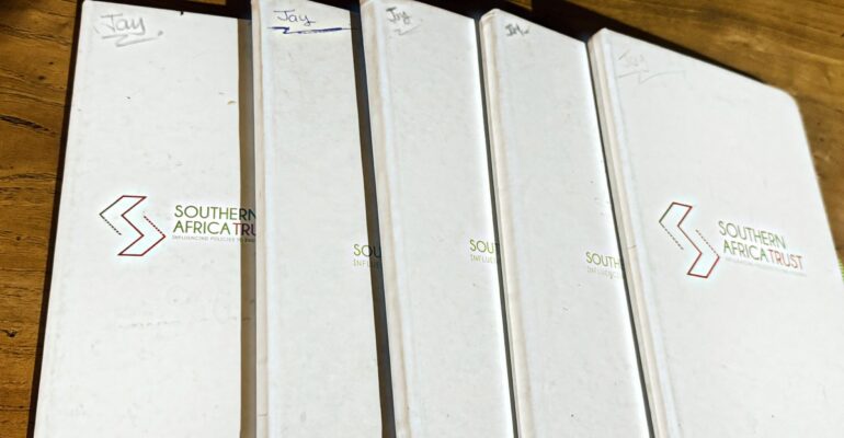 Five white Southern Africa Trust hardcover notebooks fanned out on a wooden table, each one marked "Jay" by hand in the top corner, the covers worn from years of use.