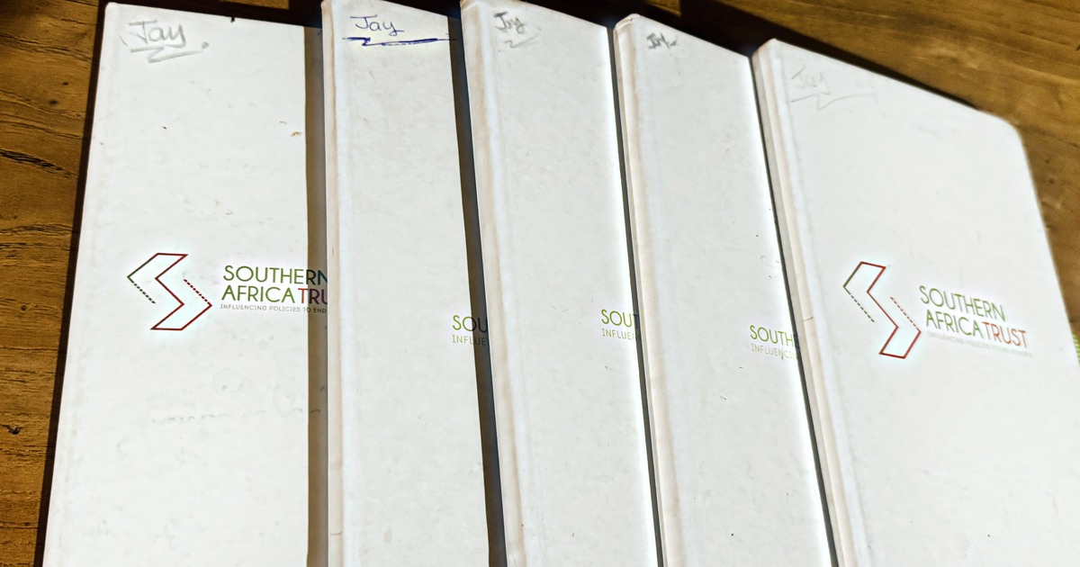 Five white Southern Africa Trust hardcover notebooks fanned out on a wooden table, each one marked "Jay" by hand in the top corner, the covers worn from years of use.